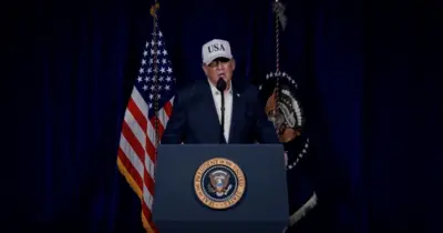 ダークスーツ姿でUSAの文字が書かれた白い野球帽をかぶったトランプ氏が、大統領印領のついた演台に向かい、マイクを前に演説している。背後には米国旗などがある